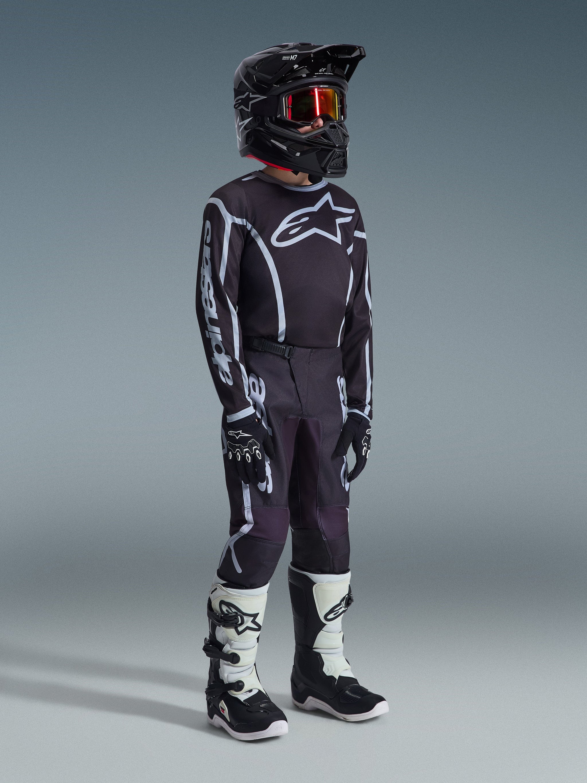 Alpinestars 2026 Youth Fluid Apex Pants, pantalon de motocross, noir et gris, porté avec un maillot noir et blanc assorti, un casque noir et des bottes blanches et noires, arborant un logo Alpinestars audacieux et des lignes graphiques, tenue de course coordonnée pour le motocross junior