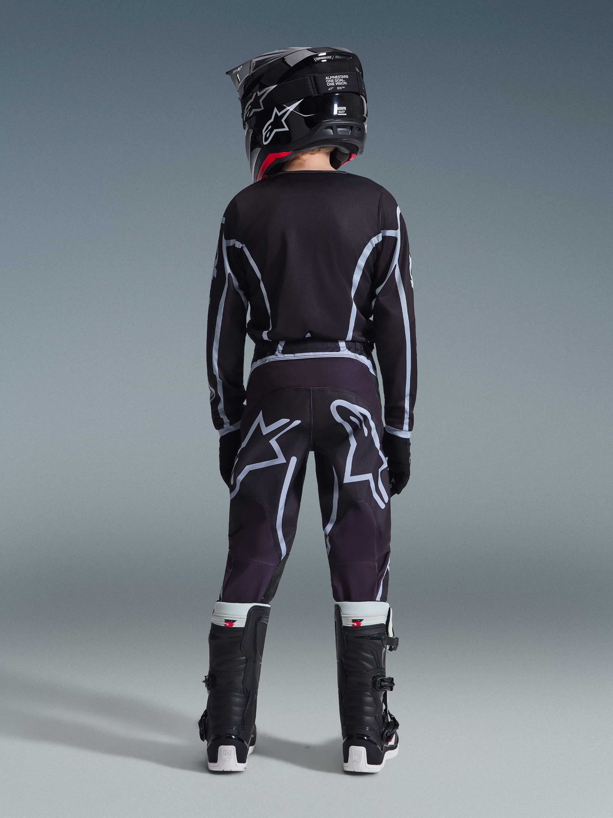 Vue arrière d'un jeune pilote de motocross portant le pantalon Alpinestars Youth Fluid Apex 2026 en noir et gris, maillot à manches longues assorti, casque noir et bottes de motocross noires et blanches, équipement de course tout-terrain coordonné.