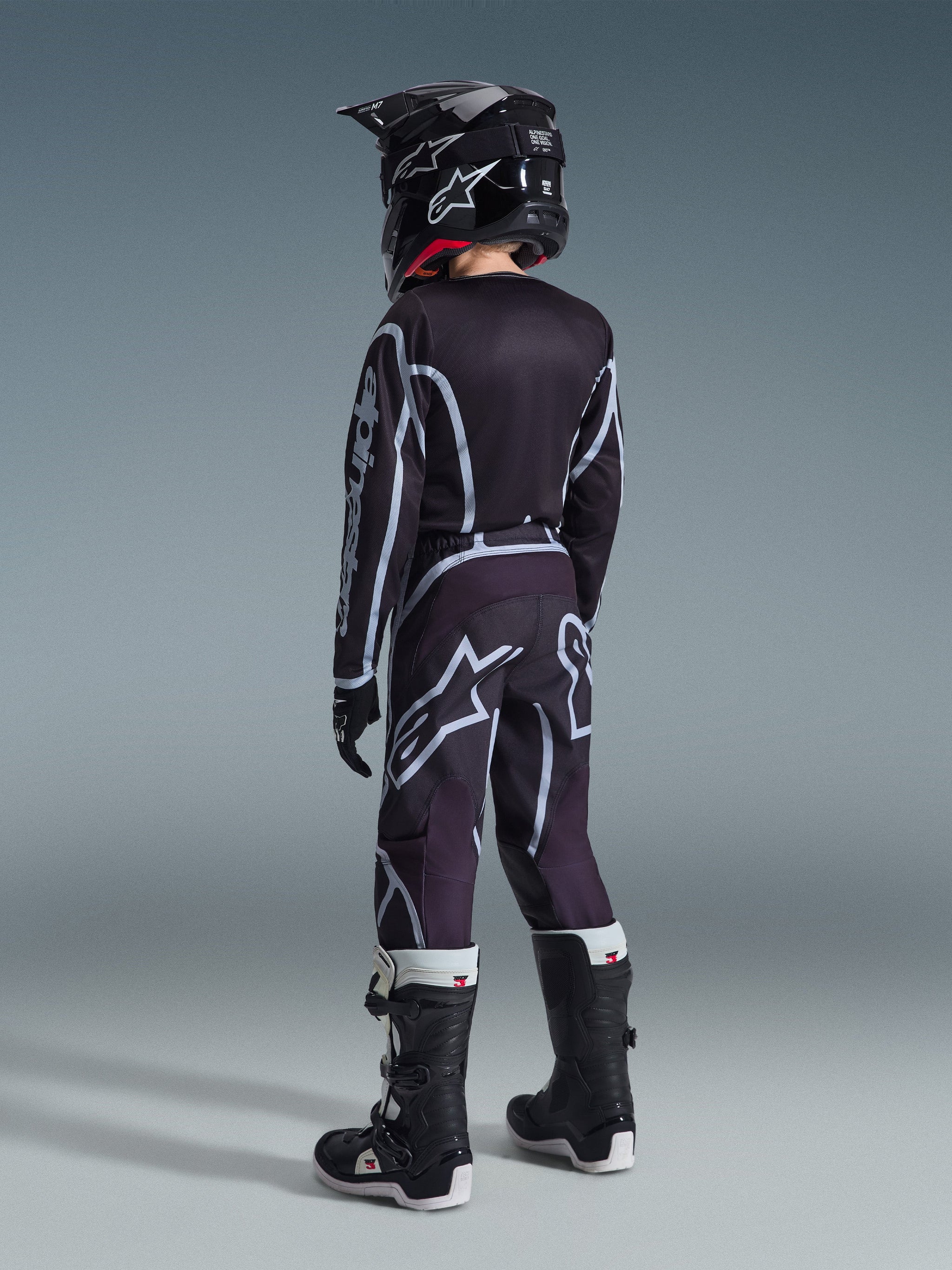 Alpinestars 2026 Youth Fluid Apex Pants, pantalon de motocross pour enfant, noir et gris, porté de dos par un jeune pilote avec un maillot et un casque noirs assortis, arborant des logos blancs imposants et des motifs géométriques, associé à des bottes noires et blanches pour la course tout-terrain.