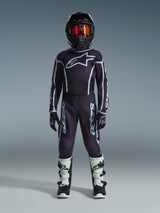 Jeune pilote de motocross Alpinestars en tenue de course complète, pantalon Youth Fluid Apex 2026 noir et gris, maillot noir assorti avec logo Alpinestars blanc, casque noir avec lunettes à effet miroir rouges, bottes blanches et noires, gants noirs et blancs, tenue coordonnée pour le tout-terrain