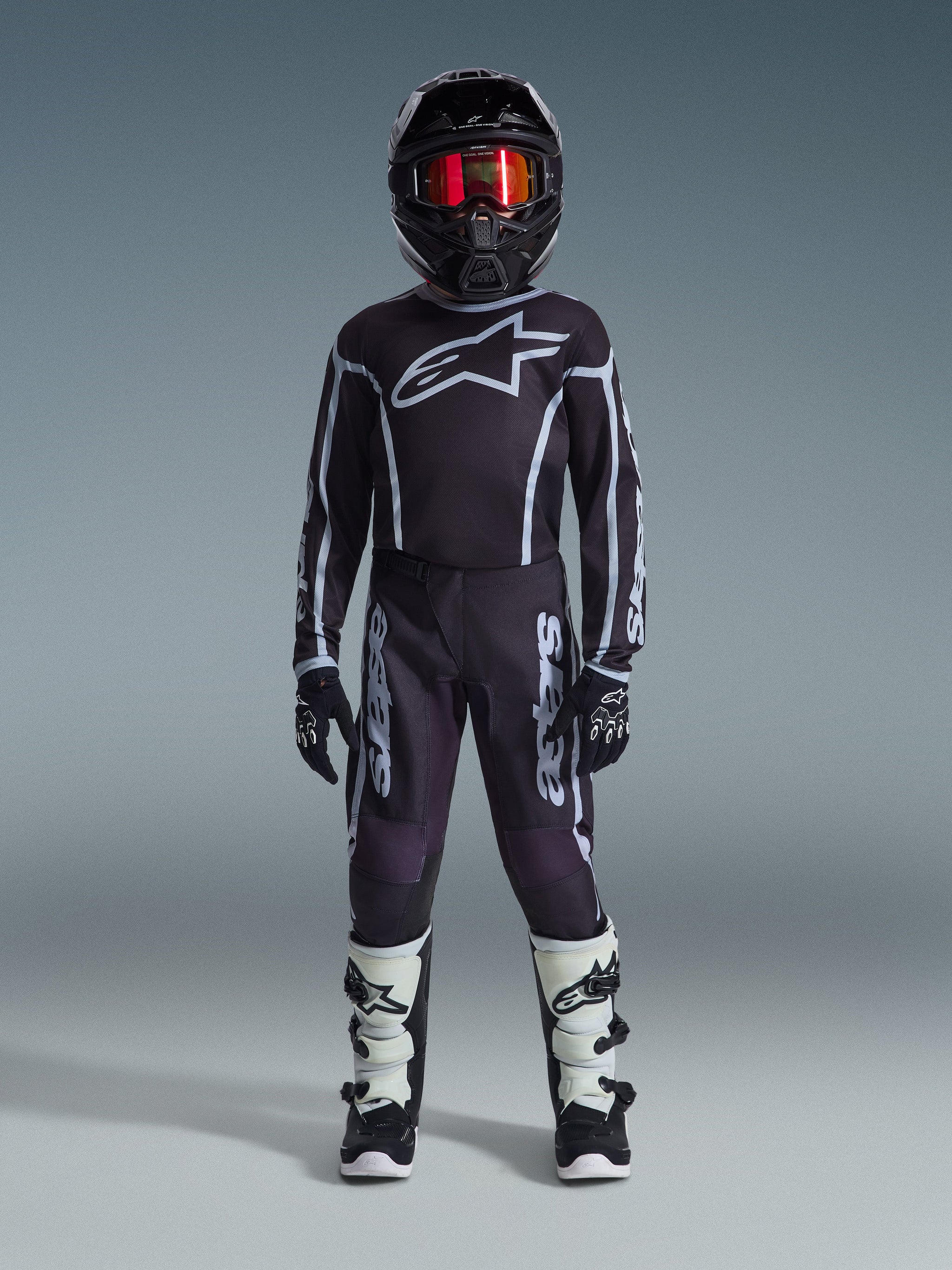 Jeune pilote de motocross Alpinestars en tenue de course complète, pantalon Youth Fluid Apex 2026 noir et gris, maillot noir assorti avec logo Alpinestars blanc, casque noir avec lunettes à effet miroir rouges, bottes blanches et noires, gants noirs et blancs, tenue coordonnée pour le tout-terrain