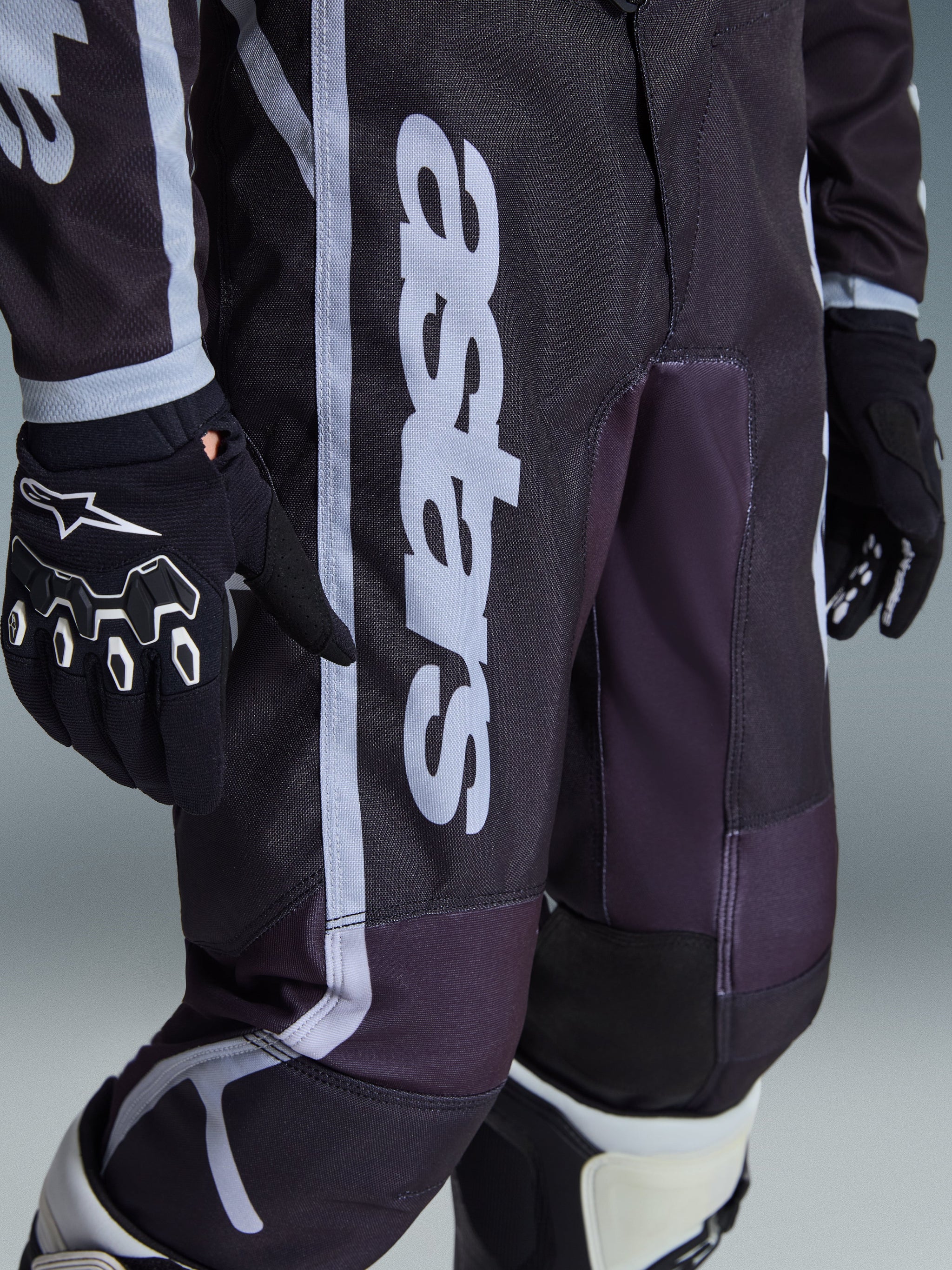 Pantalon Alpinestars Youth Fluid Apex 2026, pantalon de motocross, noir et gris, vue en gros plan montrant le logo 'astars' blanc sur la jambe, porté avec des gants et des bottes assortis noir et blanc, design sportif pour les jeunes pilotes de tout-terrain