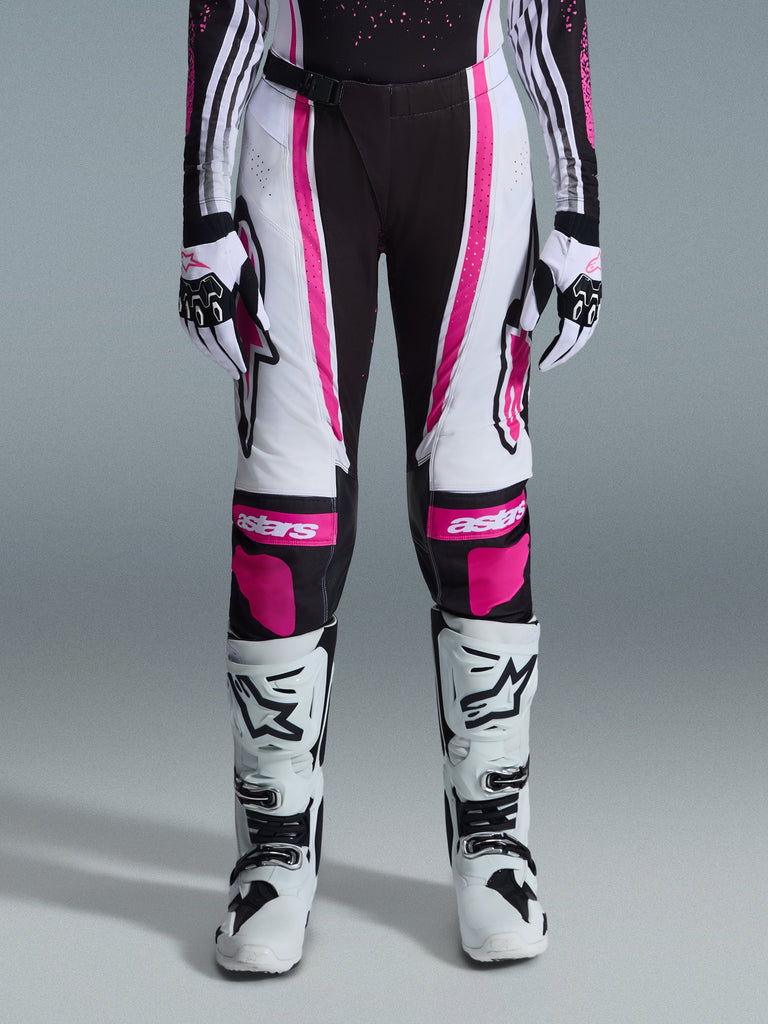 Pantalon Alpinestars Stella Techstar Nomur 2026 pour femme, pantalon de motocross, noir, blanc et fuchsia, porté avec un maillot de course, des gants assortis et des bottes de motocross blanches, doté de graphismes roses vifs et de logos astars sur les genoux, conçu pour la compétition tout-terrain de haut niveau.