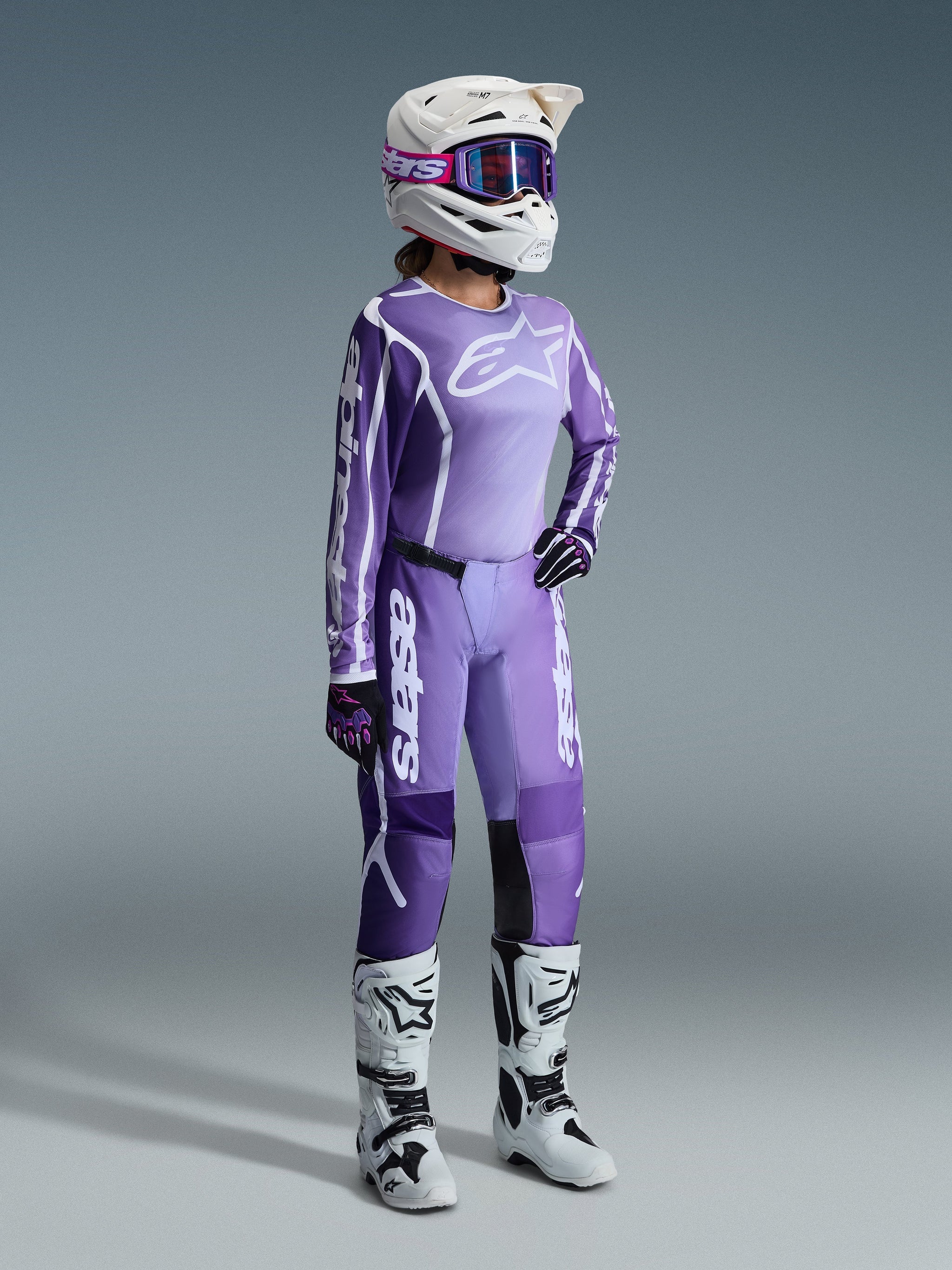 Motocross rider wearing complete racing gear, Alpinestars 2026 Stella Fluid Apex Pants in Violet and White, matching jersey with white branding, white helmet with purple goggles, white and black boots, coordinated women's outfit for off-road racing