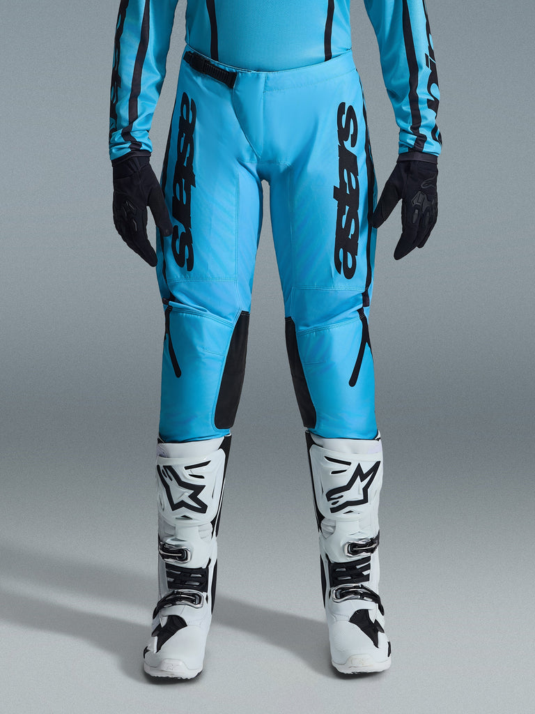 Pantalon de motocross Alpinestars Stella Fluid Apex 2026 pour femme, cyan vif et noir, logo noir vertical sur les cuisses, panneaux intérieurs de genoux renforcés noirs, présenté avec des bottes de motocross blanches et noires et des gants noirs, équipement tout-terrain professionnel pour femmes