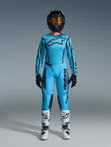 Pilote de motocross féminine portant l'ensemble Alpinestars Stella Fluid Apex 2026, maillot et pantalon bleu cyan vif avec accents noirs, casque noir avec lunettes irisées et bottes blanches, tenue de motocross professionnelle complète