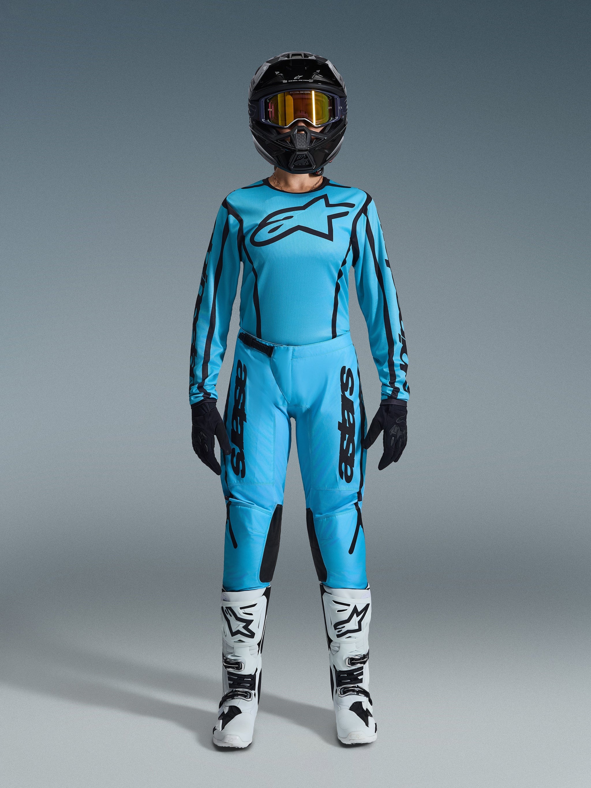 Pilote de motocross féminine portant l'ensemble Alpinestars Stella Fluid Apex 2026, maillot et pantalon bleu cyan vif avec accents noirs, casque noir avec lunettes irisées et bottes blanches, tenue de motocross professionnelle complète