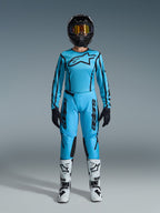 Ensemble Alpinestars 2026 Stella Fluid Apex, maillot et pantalon de motocross pour femme, bleu et noir, avec un grand logo étoile sur la poitrine, porté avec un casque noir, des lunettes teintées et des bottes blanches avec détails noirs, tenue de course tout-terrain complète