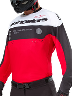 Maillot Alpinestars Pro-Dura, maillot de motocross et d'enduro, noir, rouge vif et blanc, avec le graphisme « ONE GOAL. ONE VISION. » et des perforations laser sur la poitrine, porté avec un pantalon de motocross et un casque noirs.