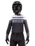 Pilote de motocross portant le maillot Alpinestars Pro-Dura en Black Magnet White, haut de performance à manches longues avec blocs de couleurs blancs et gris, porté avec un casque et des lunettes noirs, arborant le marquage « alpinestars » et le slogan « ONE GOAL ONE VISION », équipement de course de moto tout-terrain