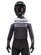 Pilote de motocross portant le maillot Alpinestars Pro-Dura en Black Magnet White, haut de performance à manches longues avec blocs de couleurs blancs et gris, porté avec un casque et des lunettes noirs, arborant le marquage « alpinestars » et le slogan « ONE GOAL ONE VISION », équipement de course de moto tout-terrain