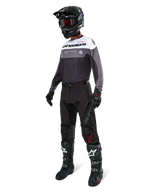 Maillot Alpinestars Pro-Dura en Black Magnet White, porté par un pilote de motocross en tenue complète avec un pantalon noir, un casque noir avec lunettes et des bottes noires et blanches. Le maillot présente un design technique color-block avec des panneaux blancs, gris et noirs, conçu pour la performance en enduro et en tout-terrain.
