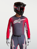Maillot Alpinestars Supertech Hiten AFD, maillot de motocross pour homme, gris et rouge fluo, mesh ventilé avec perforations au laser, porté par un pilote avec casque et gants.