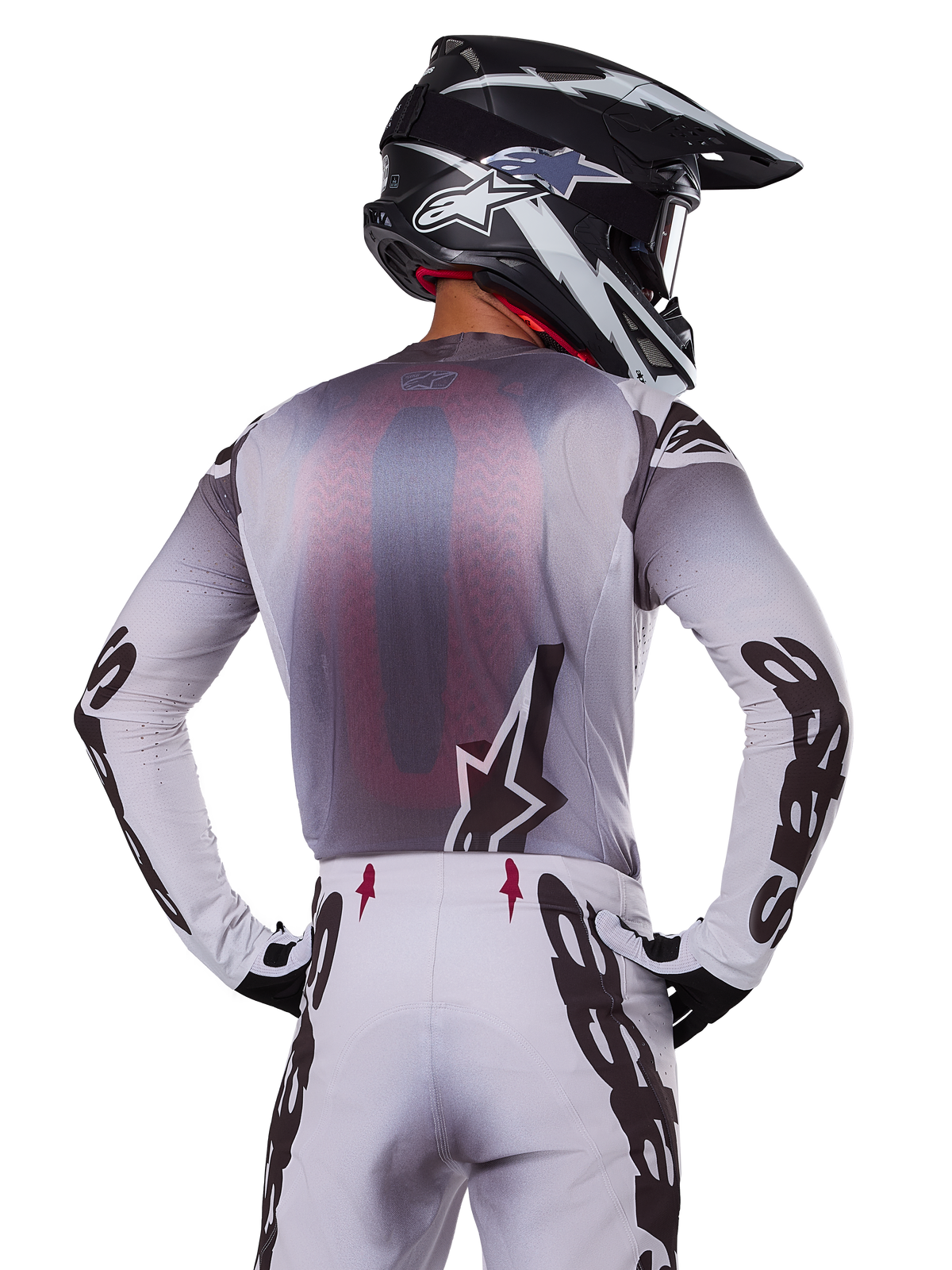 Pilote de motocross vu de dos portant le maillot Alpinestars 2025 Supertech Maker, argent et noir avec un panneau arrière en mesh rouge et des logos noirs sur les manches, associé à un pantalon de motocross assorti et un casque noir et blanc, équipement de course tout-terrain professionnel