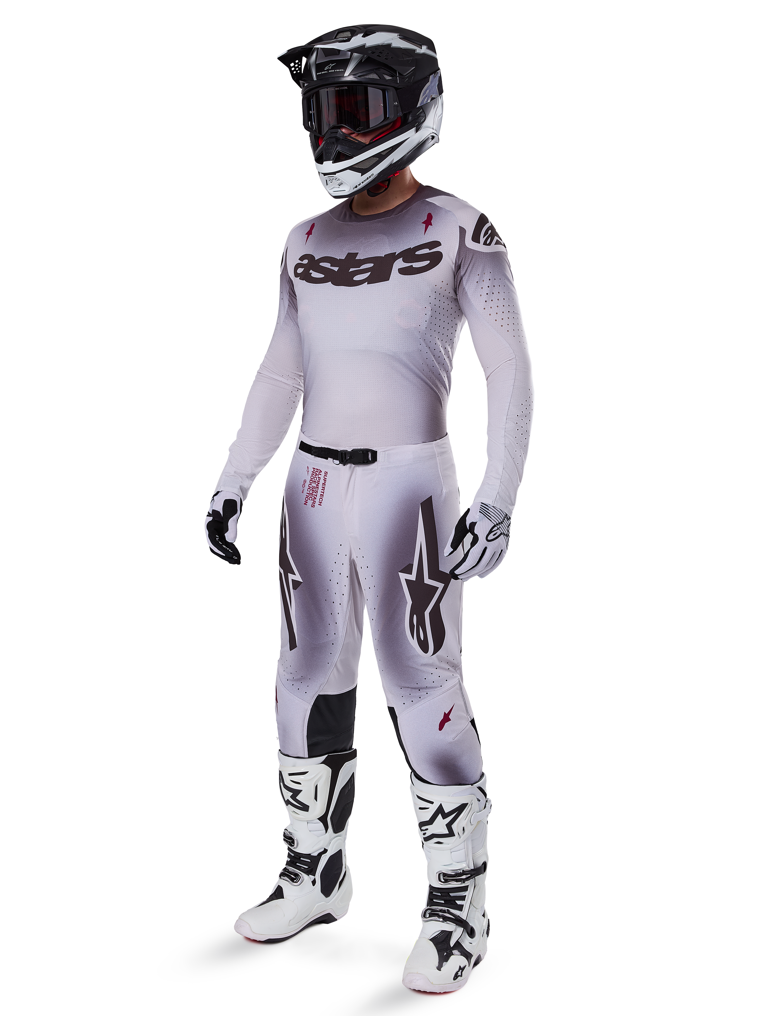 Pilote de motocross portant le maillot Alpinestars 2025 Supertech Maker en argent et noir, pantalon assorti argent et noir, casque noir et blanc avec lunettes noires, bottes blanches et noires et gants coordonnés, design dégradé professionnel pour la course tout-terrain