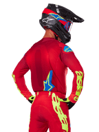 Vue arrière d'un pilote de motocross portant l'ensemble Alpinestars 2025 Supertech Maker rouge, comprenant un maillot rouge avec marquage jaune fluo sur les manches, un pantalon rouge assorti, un casque noir avec des accents bleus et rouges, et des gants noirs et jaunes coordonnés.