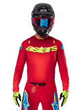 Alpinestars 2025 Supertech Maker Jersey en rouge vif et jaune fluo, avec logo 'astars' jaune sur la poitrine et motifs d'étoiles bleues sur les épaules, porté avec un pantalon assorti, un casque noir, des lunettes bleues et des gants jaunes, équipement de motocross professionnel