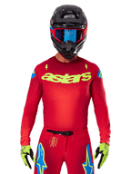 Alpinestars 2025 Supertech Maker Jersey en rouge vif et jaune fluo, avec logo 'astars' jaune sur la poitrine et motifs d'étoiles bleues sur les épaules, porté avec un pantalon assorti, un casque noir, des lunettes bleues et des gants jaunes, équipement de motocross professionnel