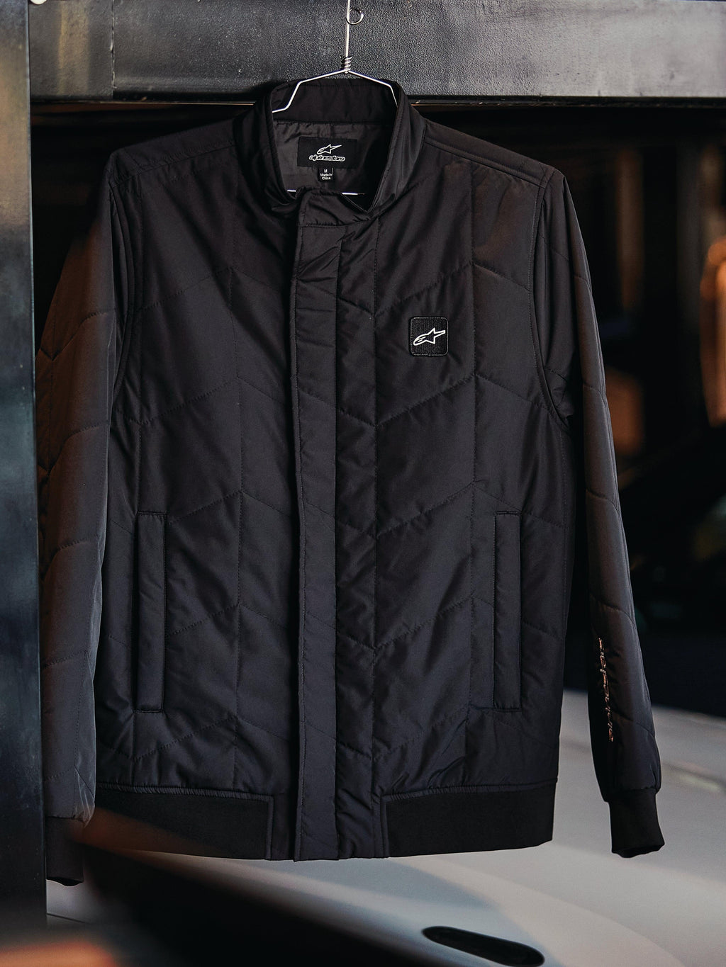 Veste Alpinestars Precedent, veste sportswear matelassée, noire, avec motif matelassé en angle, col bas et fermeture éclair couverte, écusson logo sur la poitrine, poignets et ourlet côtelés, design inspiré du sport automobile