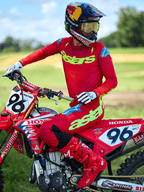 Pilote de motocross portant le maillot Alpinestars 2025 Supertech Maker, rouge vif avec logo « astars » jaune fluo, pantalon et bottes rouges assortis, gants blancs et noirs, assis sur une moto de course Honda rouge en extérieur
