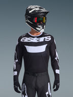 Pilote de motocross portant l'ensemble d'équipement Alpinestars Racer Air Riway 2026, maillot noir avec logo 'astars' blanc et panneau de poitrine ventilé, pantalon noir avec accents blancs, casque noir et blanc assorti avec lunettes irisées, gants noirs, équipement de motocross haute performance noir et blanc.