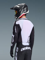 Motocross rider from behind wearing the Alpinestars 2026 Racer Air Riway Jersey, Black and White, featuring a white breathable mesh back panel and black sleeves with 'astars' branding, matching black pants with white logos, black helmet with white accents, professional off-road gear for high-temperature performance.
