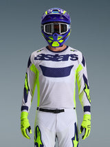 Maillot Alpinestars Racer Air Riway 2026, gris clair avec logo astars bleu foncé, accents jaune fluo et bleus sur les manches, porté par un pilote de motocross avec casque et gants assortis violet et jaune fluo, tissu en maille technique pour la course tout-terrain
