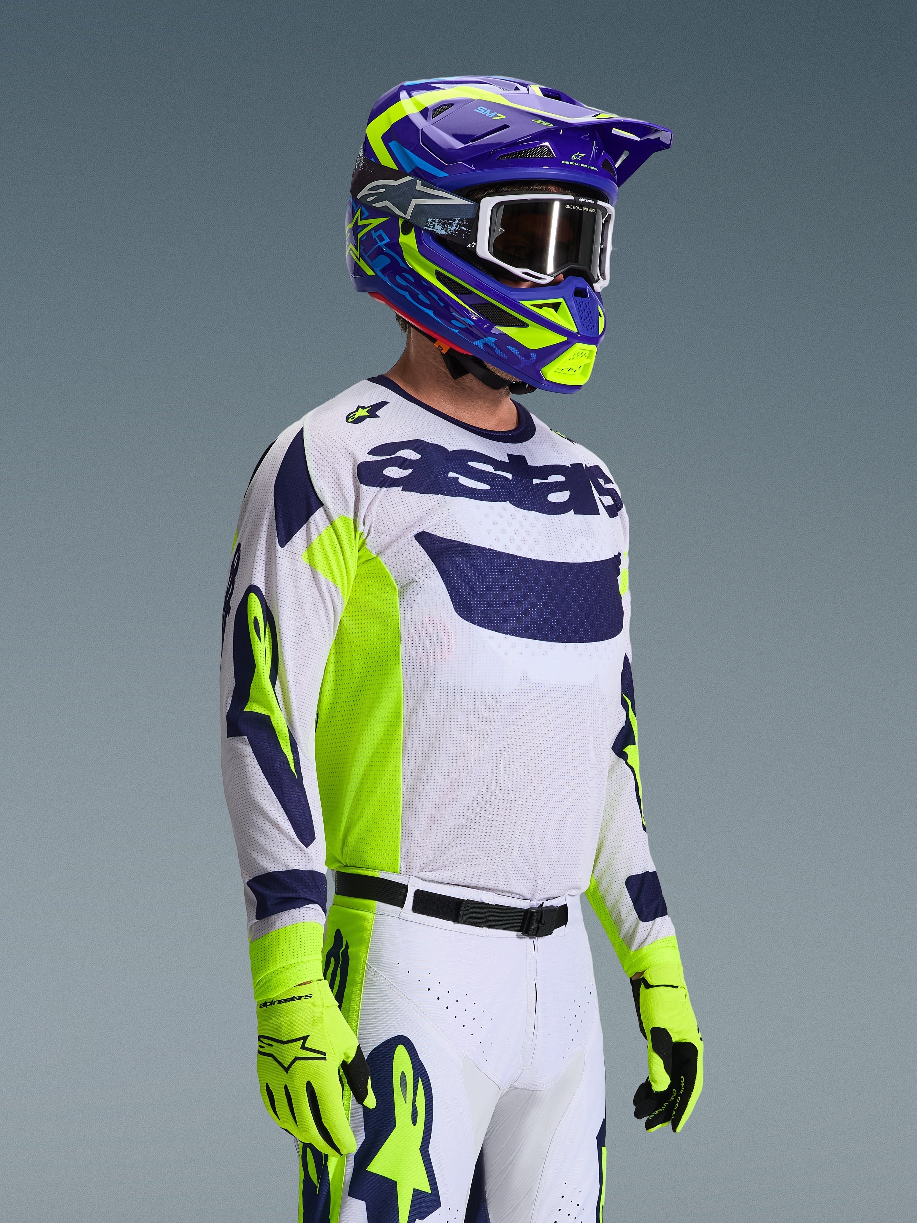 Motocross rider wearing the Alpinestars 2026 Racer Air Riway Gear Set, white jersey with navy blue logo and neon yellow side panels, white pants with matching navy and yellow accents, purple and neon yellow helmet with white goggles, neon yellow gloves, high-performance off-road racing gear