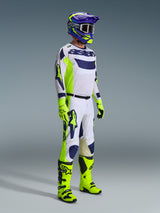Pilote de motocross Alpinestars portant une tenue de course complète, maillot gris clair avec des accents jaune fluo et bleus, pantalon assorti blanc et jaune, bottes et gants jaune vif, casque violet et jaune avec lunettes foncées, tenue coordonnée pour la course tout-terrain