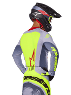 Maillot Alpinestars Techstar Melt 2025, Rouge Vif Gris, vue arrière d'un pilote de motocross portant une tenue de course grise et jaune fluo, logo étoile rouge dans le dos, casque noir et rouge assorti, équipement tout-terrain haute performance