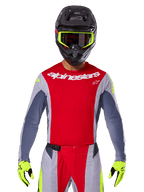 Pilote de motocross portant le maillot Techstar Melt 2025 et le pantalon assorti, rouge vif et gris avec logo Alpinestars blanc, casque noir avec lunettes jaune fluo, gants jaune fluo assortis, équipement de performance ventilé pour la course tout-terrain