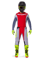 Pilote de motocross portant l'ensemble d'équipement Alpinestars Techstar Melt 2025, maillot rouge avec manches grises et logo blanc, pantalon assorti gris et rouge, lunettes, gants et bottes jaune fluo, tenue de motocross complète pour la performance tout-terrain.