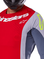 Alpinestars Techstar Melt Jersey 2025, maillot de motocross, rouge vif et gris avec empiècements bleu foncé, logos blanc et jaune fluo sur l'épaule, gros plan montrant les perforations de ventilation découpées au laser et un casque noir