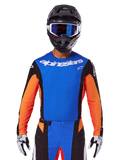 Maillot Alpinestars Techstar Melt 2025, maillot de motocross bleu et orange avec empiècements latéraux noirs, porté avec un casque noir et des lunettes bleues, doté de perforations laser respirantes sur la poitrine, coupe athlétique de compétition pour performance tout-terrain