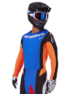 Ensemble d'équipement Alpinestars Techstar Melt 2025, pilote de motocross portant un maillot bleu avec manches orange et panneaux latéraux noirs, pantalon assorti bleu et noir, casque noir avec lunettes bleues, gants blancs et noirs, tenue de motocross à coupe sportive de compétition