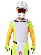 Alpinestars 2025 Techstar Melt Jersey, motocross racing gear, white chest with neon yellow and orange sleeves, rider wearing white helmet and red goggles, neon yellow gloves with black logos, vibrant colorway for off-road performance