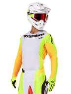 Motocross rider wearing Alpinestars 2025 Techstar Melt Gear Set, white jersey with vibrant yellow and orange sleeves, black 'Alpinestars' chest branding, matching neon yellow gloves, white helmet with red-tinted goggles, professional off-road racing kit