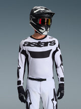 Pilote de motocross portant le maillot Alpinestars Racer Riway 2026 blanc et noir, avec le logo 'astars' sur la poitrine, associé à un pantalon blanc et noir assorti, un casque noir avec lunettes et des gants noirs, tenue coordonnée pour la course tout-terrain