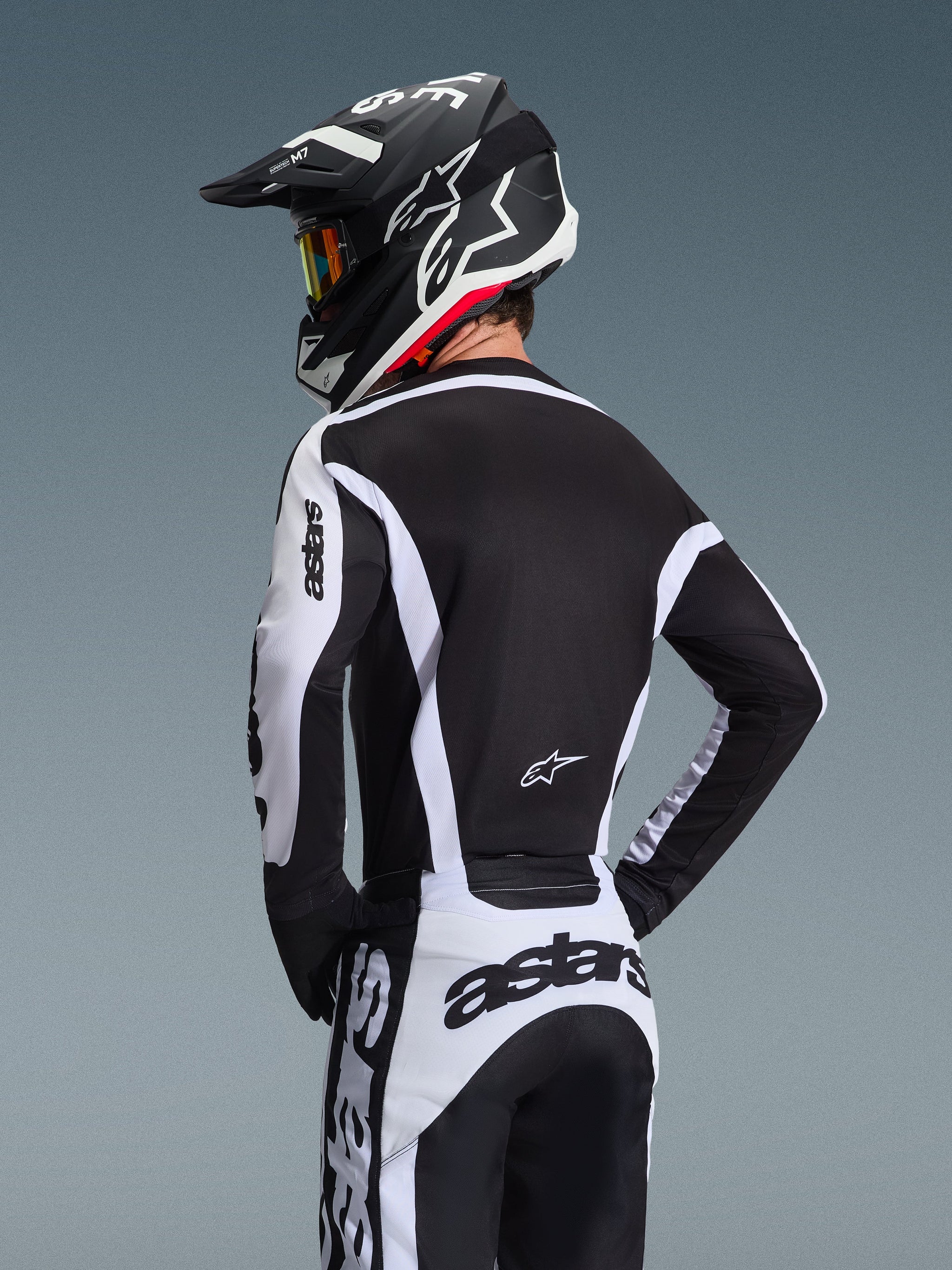 Pilote de motocross vu de dos portant le maillot Alpinestars Racer Riway 2026 blanc et noir, avec le marquage 'astars' sur les manches et le bas du dos, associé à un pantalon de motocross assorti et un casque noir avec lunettes, équipement coordonné pour la course tout-terrain.