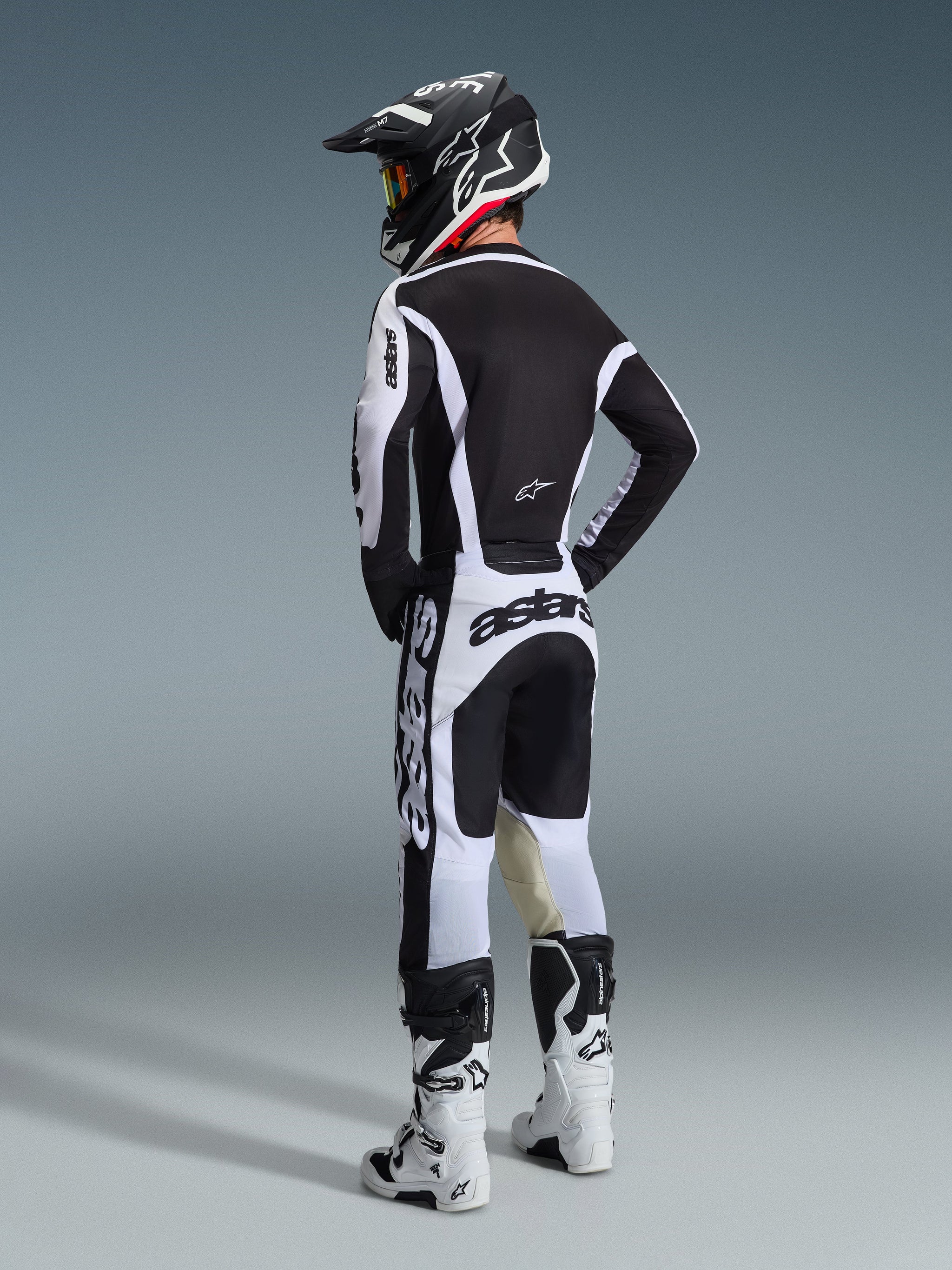 Pilote de motocross portant une tenue de course complète vue de dos, maillot Alpinestars Racer Riway 2026 blanc et noir, pantalon blanc et noir assorti avec marquage 'astars' imposant, bottes blanches et casque noir avec accents blancs, ensemble de performance coordonné pour la course tout-terrain.
