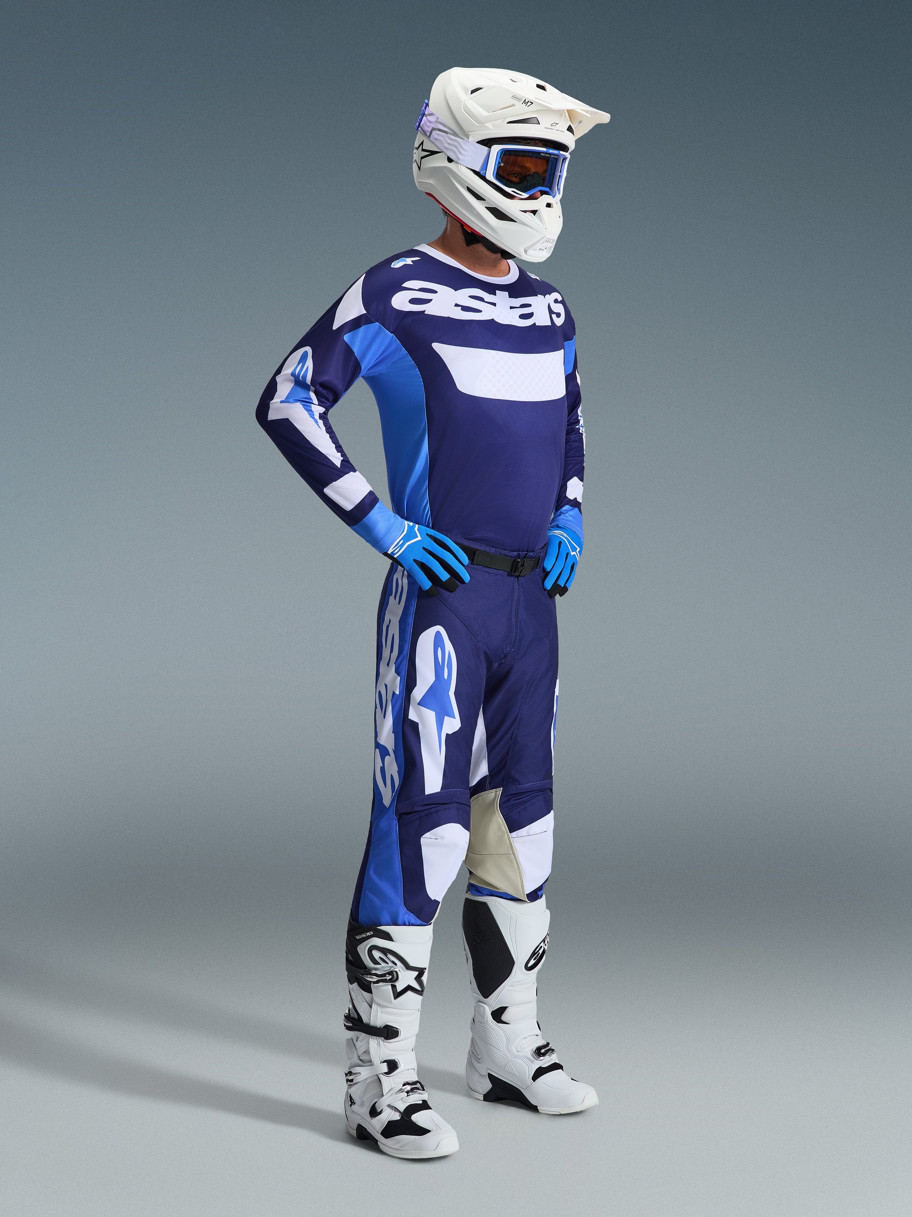 Pilote de motocross portant l'ensemble d'équipement Alpinestars Racer Riway 2026 en bleu UCLA et blanc, comprenant un maillot bleu et blanc avec logo 'astars', un pantalon assorti, un casque blanc avec lunettes bleues et des bottes tout-terrain blanches, tenue de course complète pour le motocross.