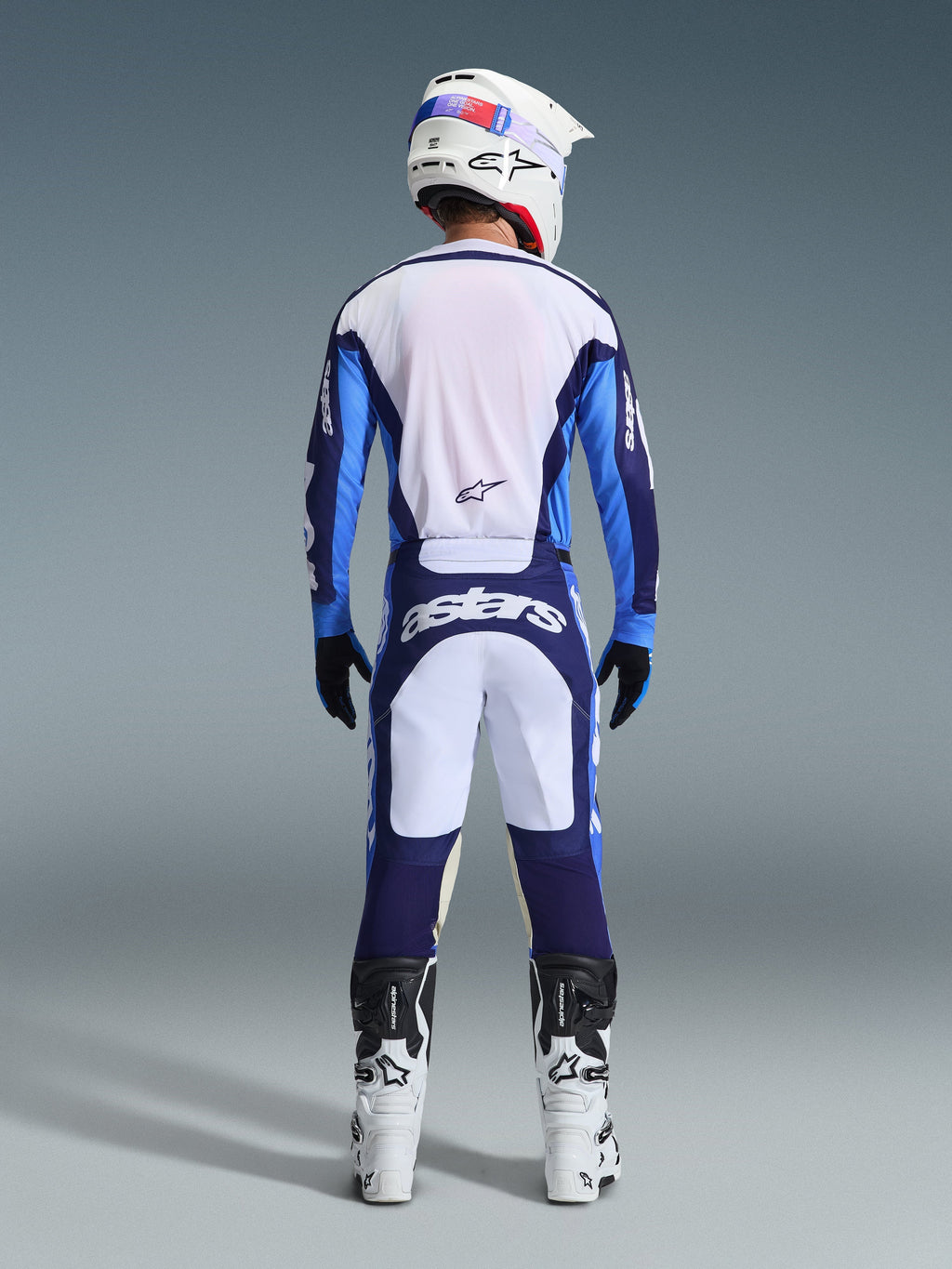 Vue arrière d'un pilote de motocross portant le maillot et le pantalon Alpinestars Racer Riway 2026 en bleu UCLA et blanc, coordonné avec un casque blanc, des gants noirs et des bottes de motocross blanches, équipement haute performance pour la course tout-terrain