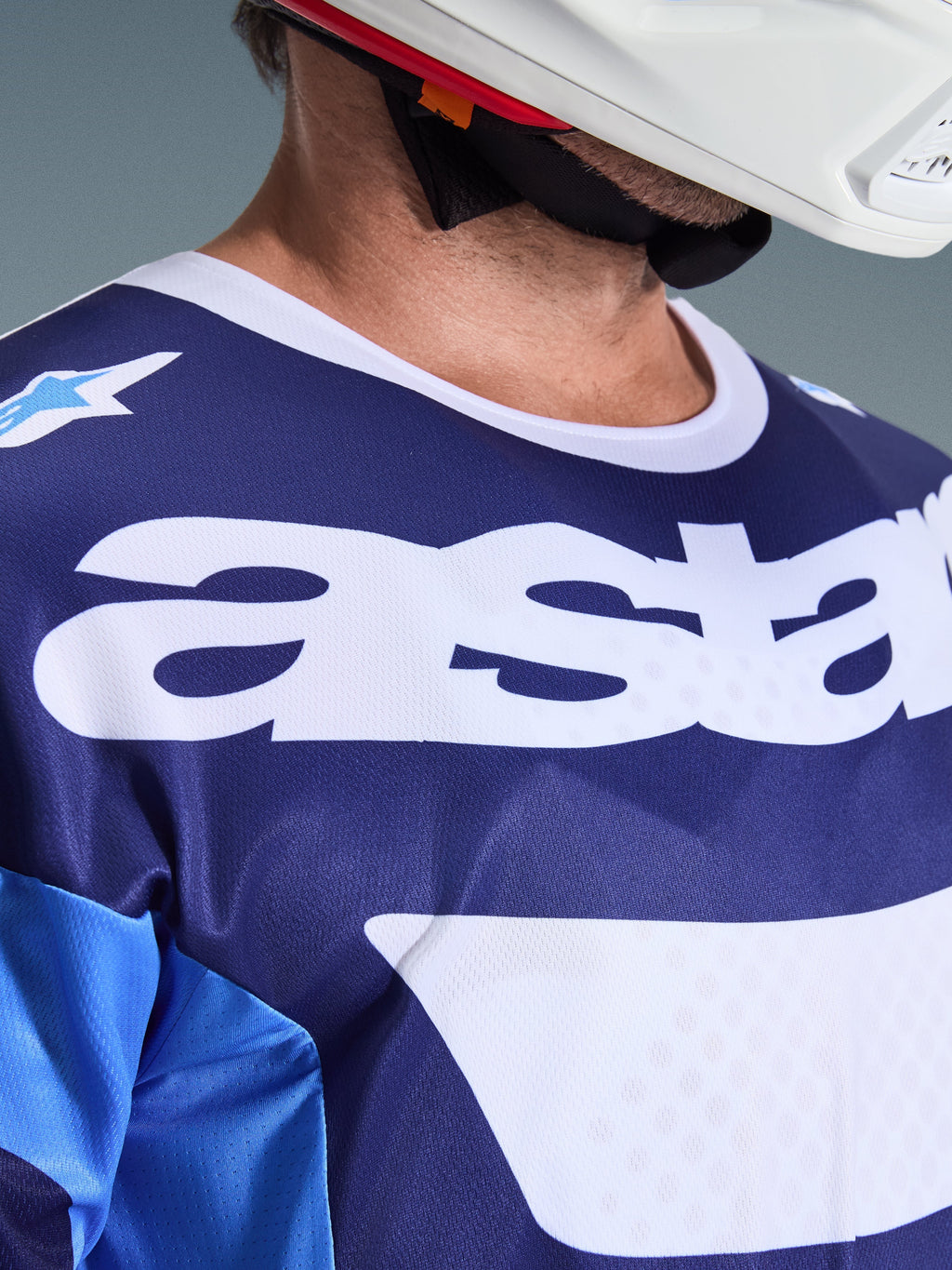 Alpinestars Racer Riway Jersey 2026, maillot de motocross, bleu UCLA et blanc, vue rapprochée de la poitrine avec grand marquage 'astars' blanc, col blanc et texture en maille respirante, porté avec un casque blanc