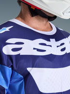 Alpinestars Racer Riway Jersey 2026, maillot de motocross, bleu UCLA et blanc, vue rapprochée de la poitrine avec grand marquage 'astars' blanc, col blanc et texture en maille respirante, porté avec un casque blanc