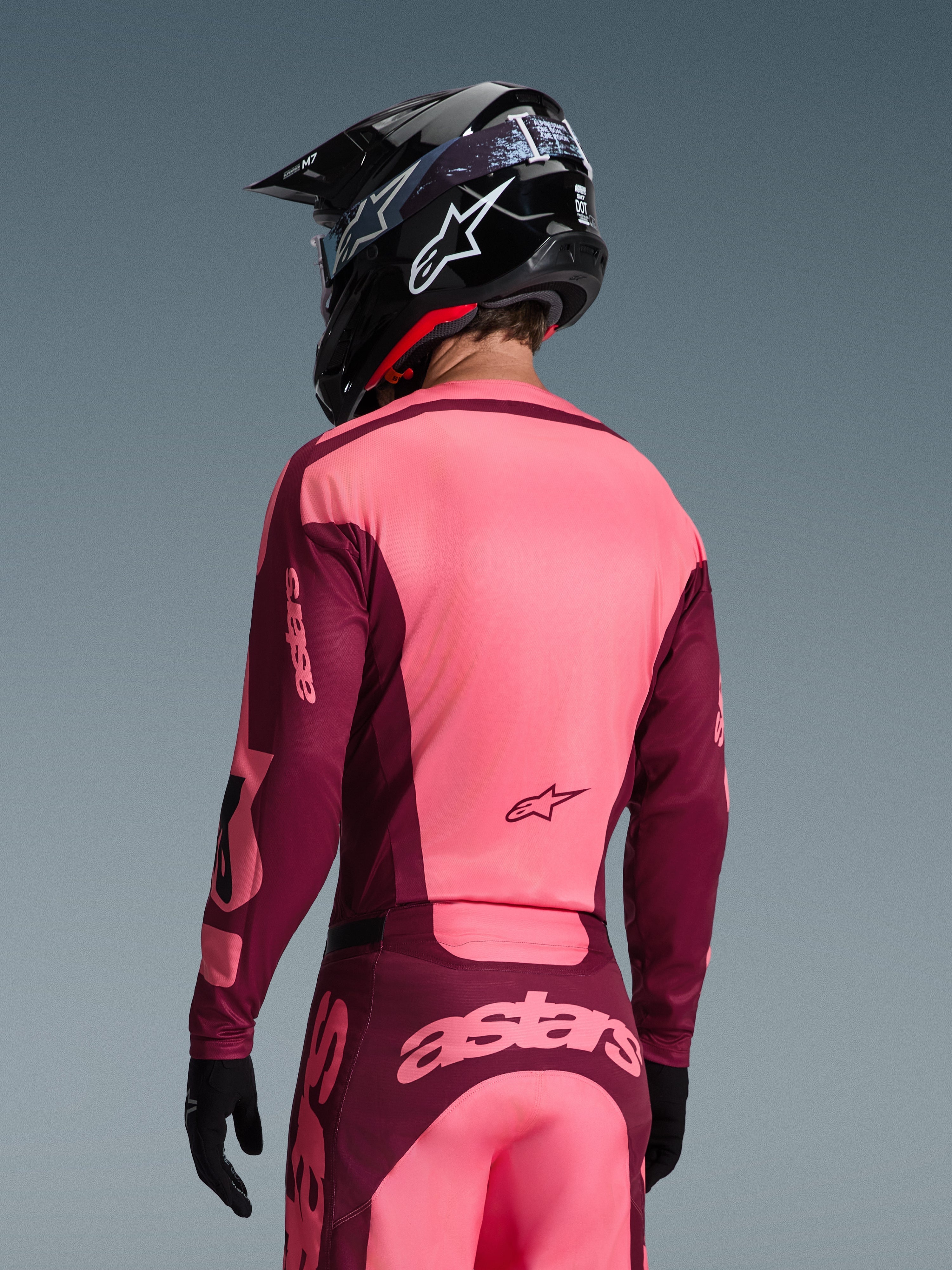 Motocross rider from behind wearing Alpinestars 2026 Racer Riway Jersey and matching pants, Dark Red and Coral colorway, black helmet with white Alpinestars logo, black gloves, coordinated professional gear for off-road racing