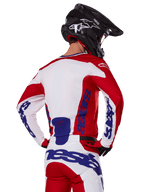 Ensemble maillot et pantalon Alpinestars Racer Veil 2025, tenue de motocross, rouge vif, blanc et bleu, vue arrière d'un pilote portant l'équipement coordonné et un casque de motocross noir, arborant le logo Alpinestars bleu, vêtement technique de course tout-terrain