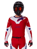 Maillot Alpinestars Racer Veil 2025, maillot de motocross, rouge, blanc et bleu, à manches longues, porté avec un casque noir et des lunettes orange, gants et pantalon rouges assortis, design performant pour la conduite tout-terrain