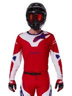 Maillot Alpinestars Racer Veil 2025, maillot de motocross, rouge, blanc et bleu, à manches longues, porté avec un casque noir et des lunettes orange, gants et pantalon rouges assortis, design performant pour la conduite tout-terrain