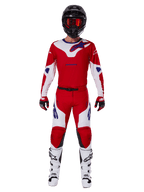 Pilote de motocross portant l'ensemble Alpinestars Racer Veil 2025, maillot et pantalon rouge vif et blanc avec accents bleus, casque noir avec lunettes rouges, bottes blanches et gants rouges et noirs, tenue coordonnée pour la course tout-terrain