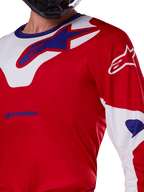 Maillot Alpinestars Racer Veil 2025, maillot de motocross, rouge vif et blanc, vue rapprochée du haut du torse et de la poitrine, logo Alpinestars bleu sur un empiècement de col blanc, manches rouges avec accents blancs, porté avec un casque noir, vêtement technique de conduite tout-terrain